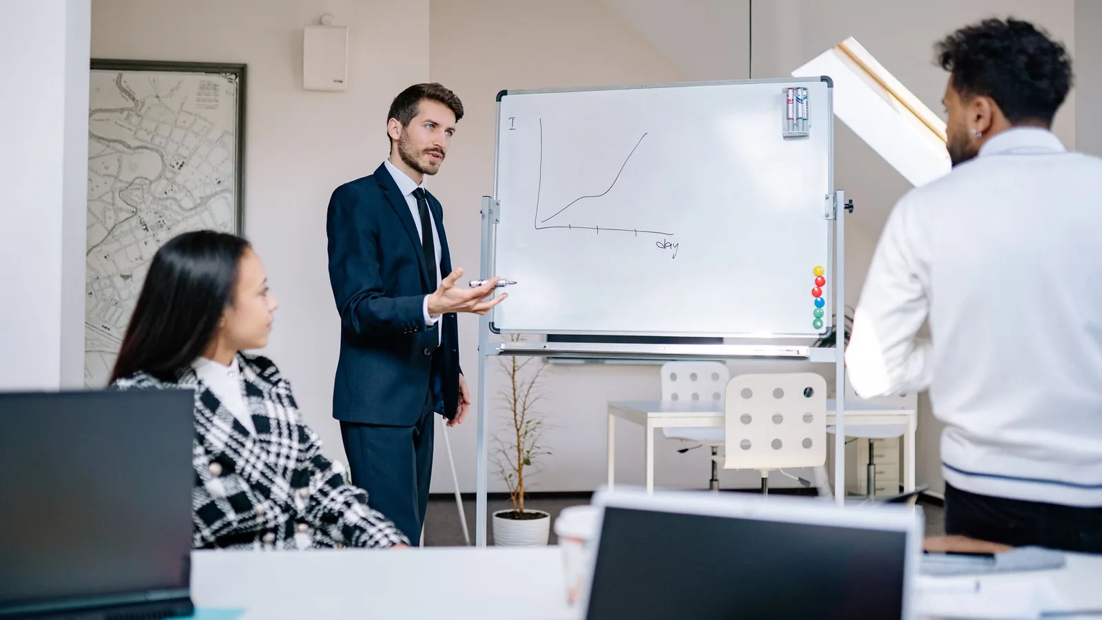 A consultant in a suit explains a growth curve on a whiteboard to two people in an office.