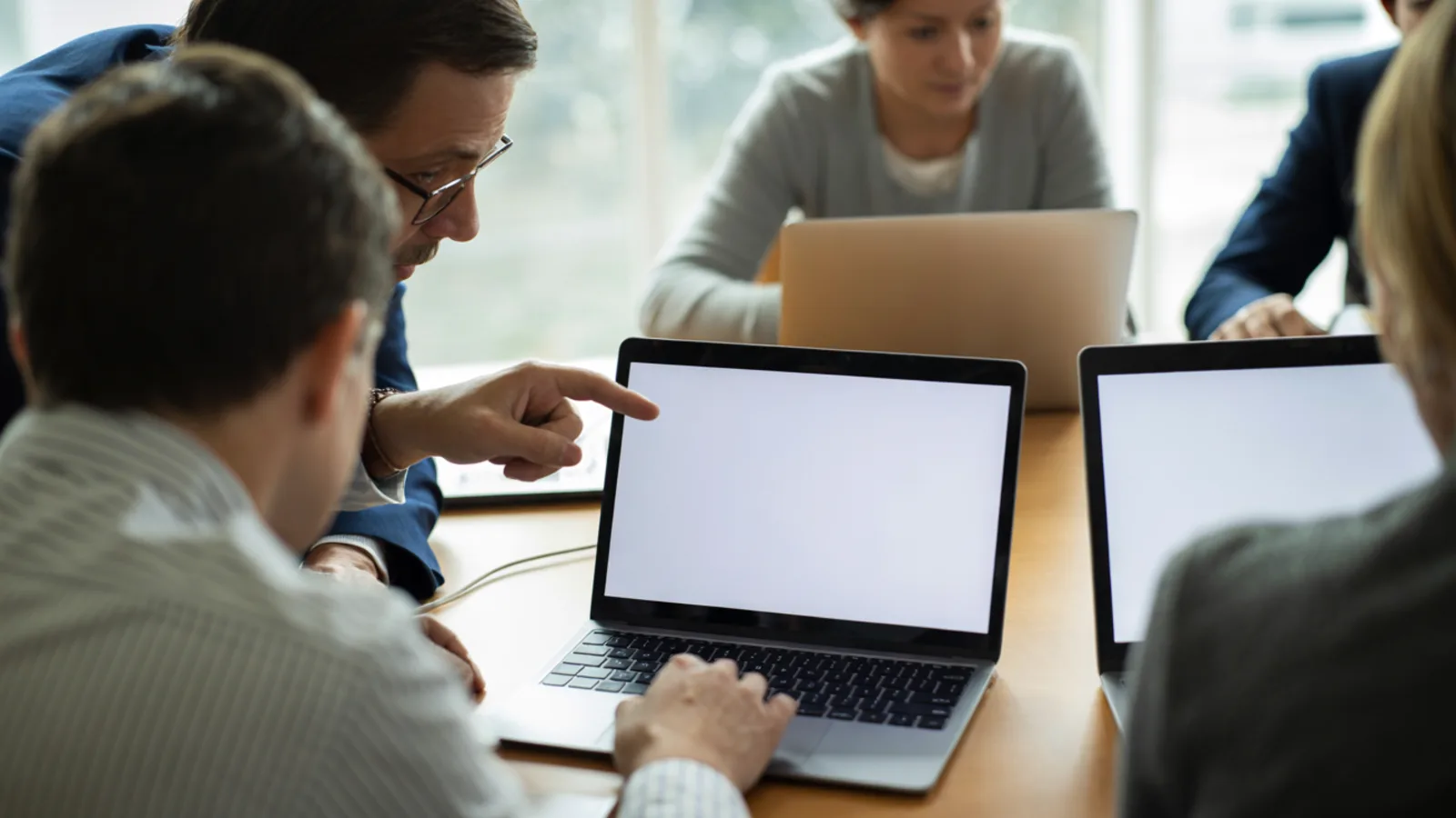 An instructor leans in beside a seated learner, pointing at their blank laptop screen, with two more learners working at their own laptops softly out of focus in the background of a bright classroom.